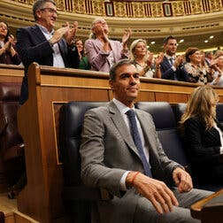 Pedro Sánchez, in a gray suit and blue tie, smiles while seated in the Spanish Parliament. Many people in the background stand clapping.