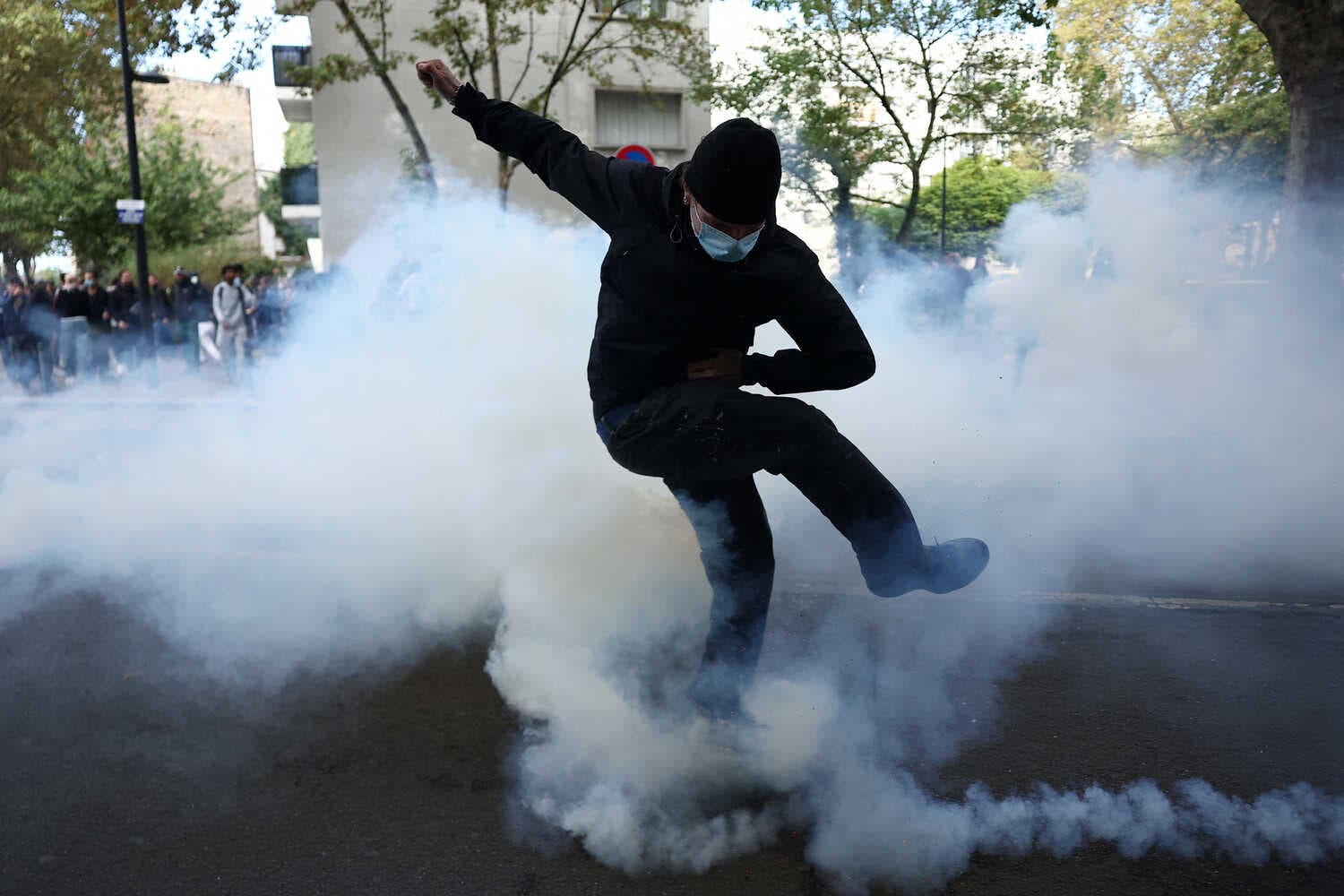 A person in a dark hooded sweatshirt and blue surgical mask kicks through a cloud of white smoke.
