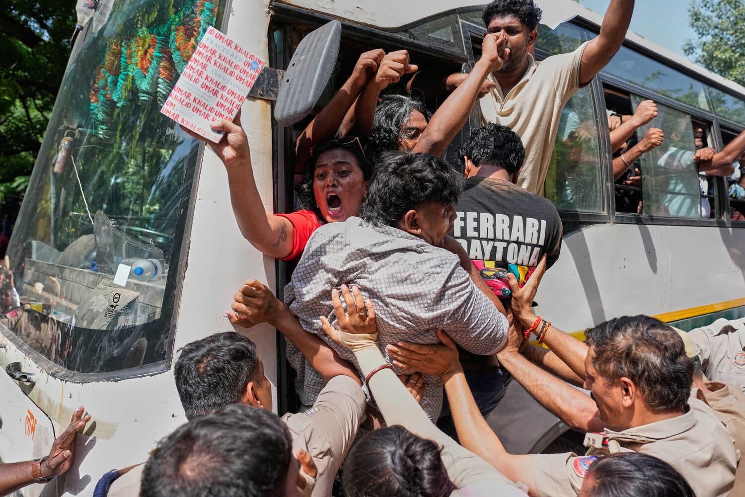 People are tightly packed and being pushed through the door of a white bus by others in uniform. A person in a red shirt, mouth open wide, holds up a book.