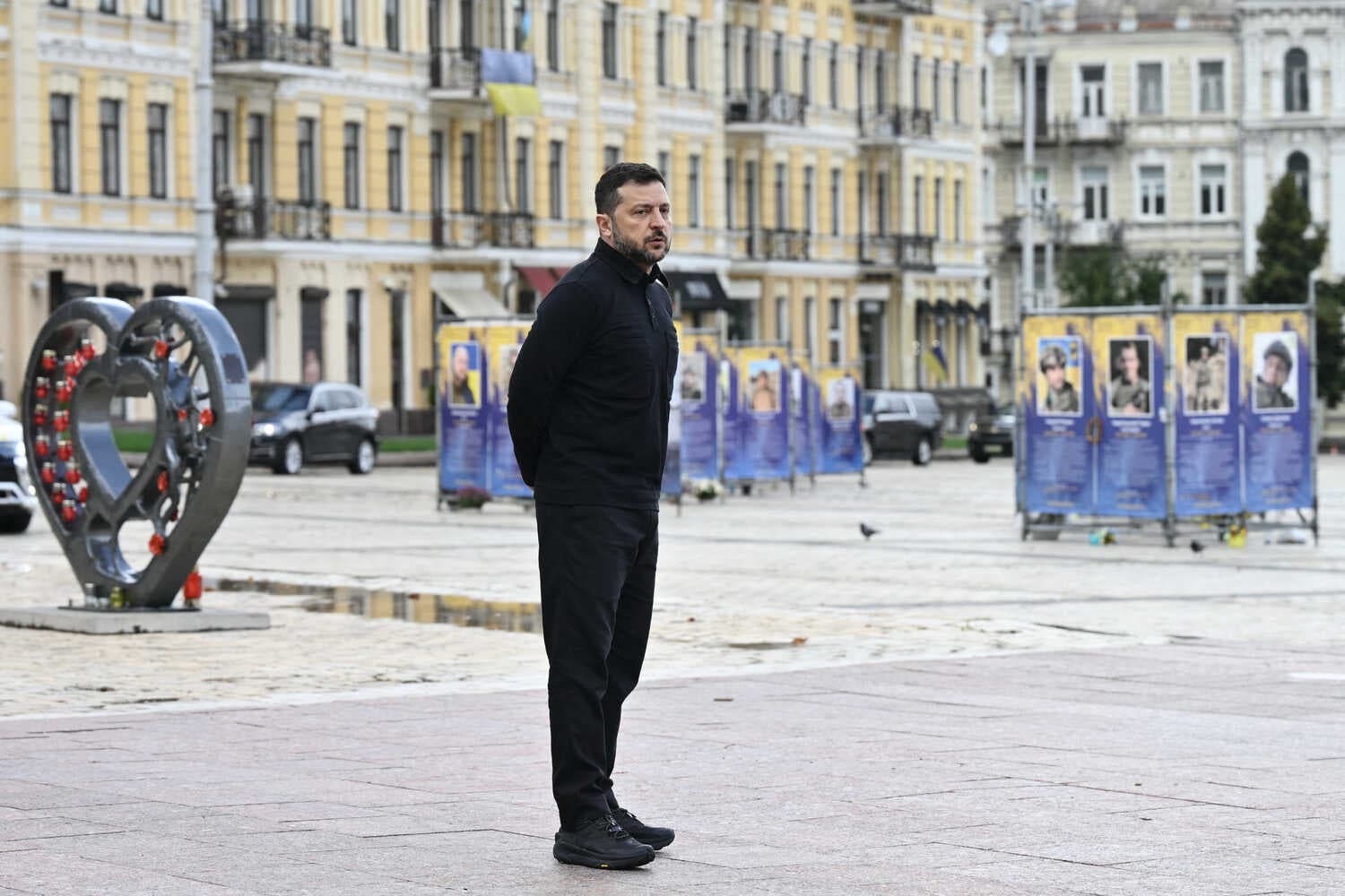 Mr. Zelensky, in black, stands on a paved square near a heart-shaped memorial. In the background are buildings and blue banners with portraits.