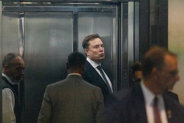 Elon Musk, wearing a suit and tie, stands next to an elevator.